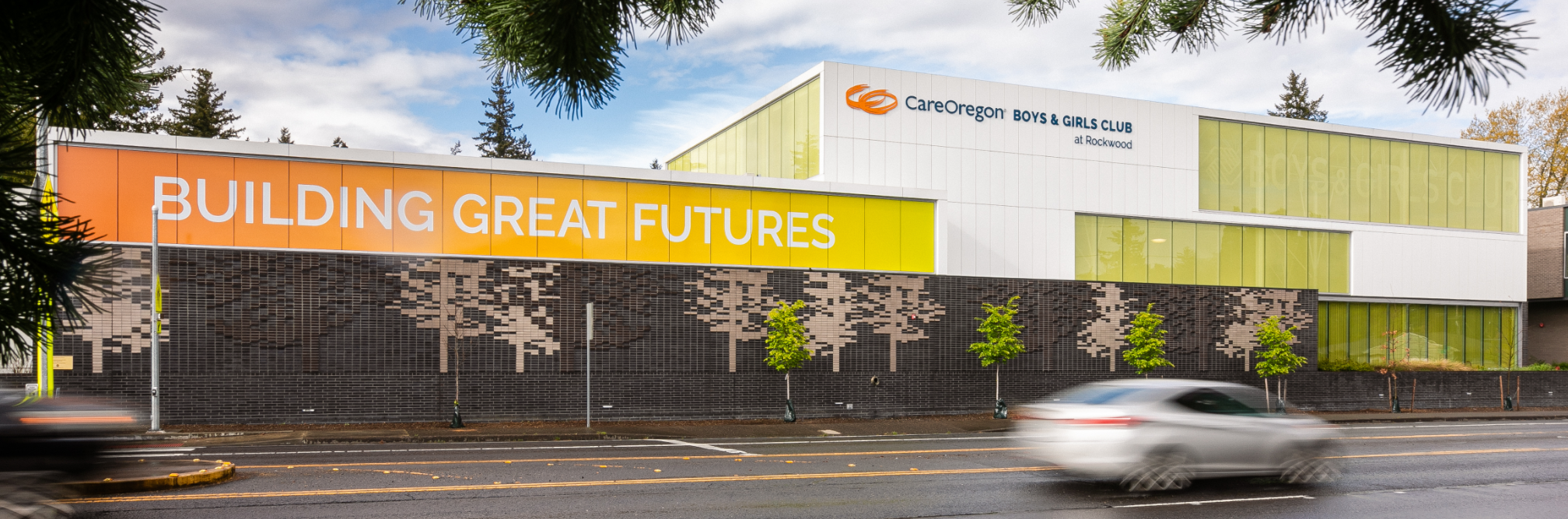 Against shades of orange, yellow and yellow-green, Building great futures, in large white capital letters, spans a white rectangular building. A sign on top of the building identifies it as CareOregon Boys & Girls Club at Rockwood.