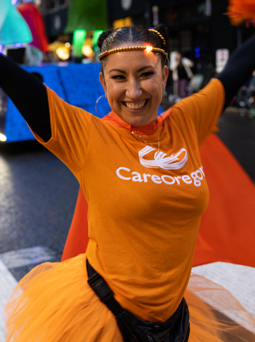 Smiling young woman wearing an orange CareOregon T-shirt and orange net tutu, happily throwing her arms in the air as she walks past a crowd of parade-goers.
