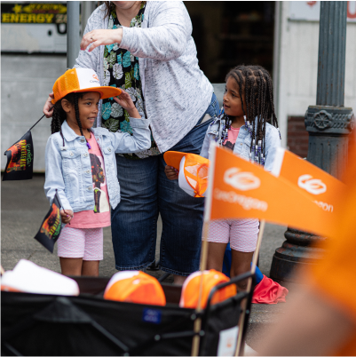 Two children receiving orange hats at an outdoor event, with an adult assisting them, in a street parade setting 