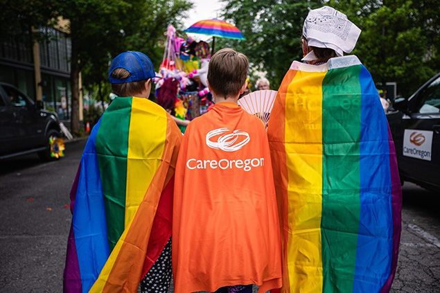 Se muestra de espaldas a tres niños, los dos de los extremos lucen capas con un diseño de arcoiris y el que se encuentra en el medio lleva una capa naranja con el logotipo de CareOregon en el centro