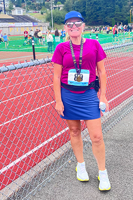 Ratina-track A woman in a fuscia shirt and blue running skirt, running shoes and sunglasses standing next to a red running track, smiling, holding a water bottle