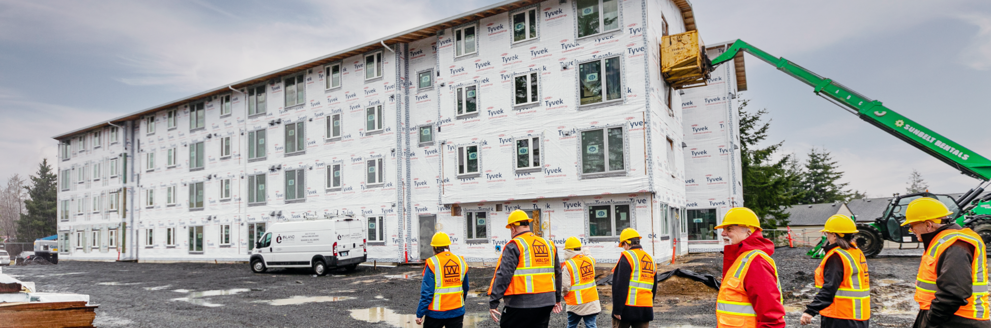 A group of seven adults in yellow hard hats and orange reflective vests approaches a four-story apartment building, covered with Tyvek, being built in Clatsop County.