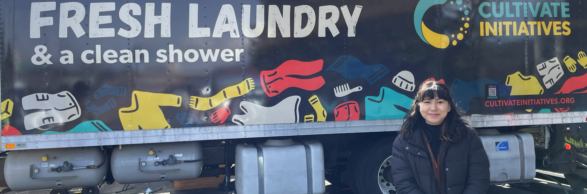 The side of a semi-truck-sized mobile shower unit is covered with cartoons of clothing, the large words, Fresh Laundry and a clean shower and the logo for Cultivate Initiatives. In front stands a female CareOregon employee who hands out hygiene kits to people using the service.