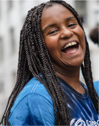 Person with braided hair wearing a blue shirt, standing in an outdoor setting with a blurred background.