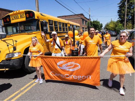 A group of cheerful people wearing orange t-shirts and tutus, holding a ‘CareOregon’ banner, as they march in a parade