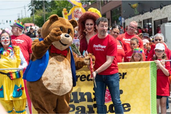 Red t-shirt-clad participants in a festive parade, holding a yellow banner, a person in a bear costume and a clown 