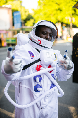 一个穿着宇航员服装的人站在城市街道上，脸被遮住，背景是模糊的树木