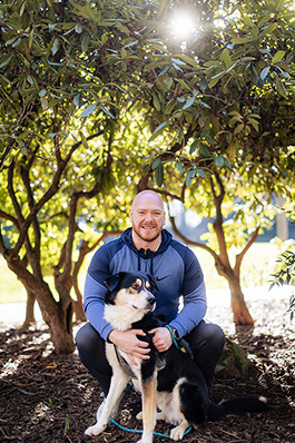 careoregon_kris_feb2025_fj6a3913_jasonquigley-story A man crouches under some trees, posing for the camera with his dog